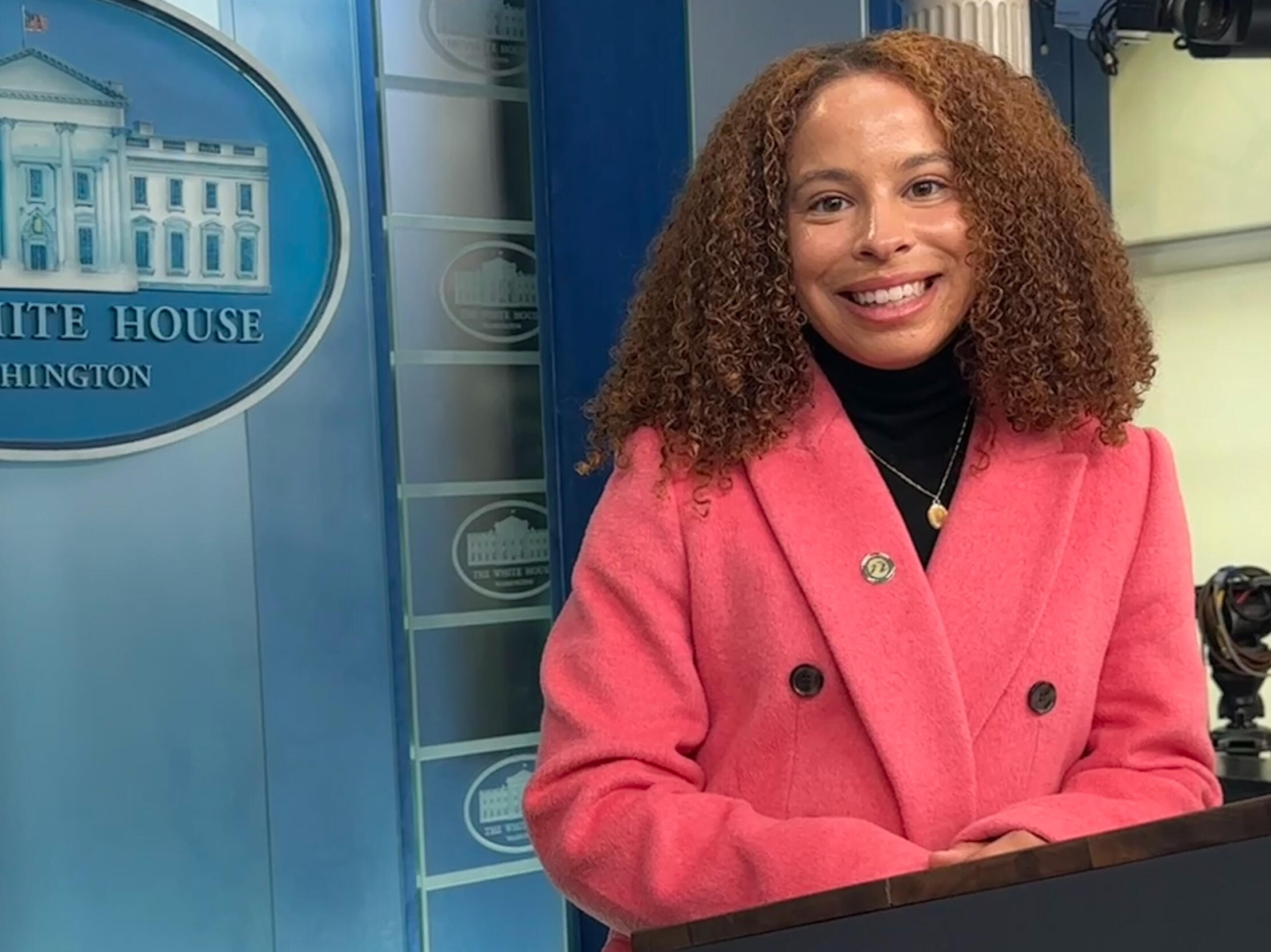 Avril Silva pictured in the White House Press Briefing room wearing a pink coat and black turtleneck.