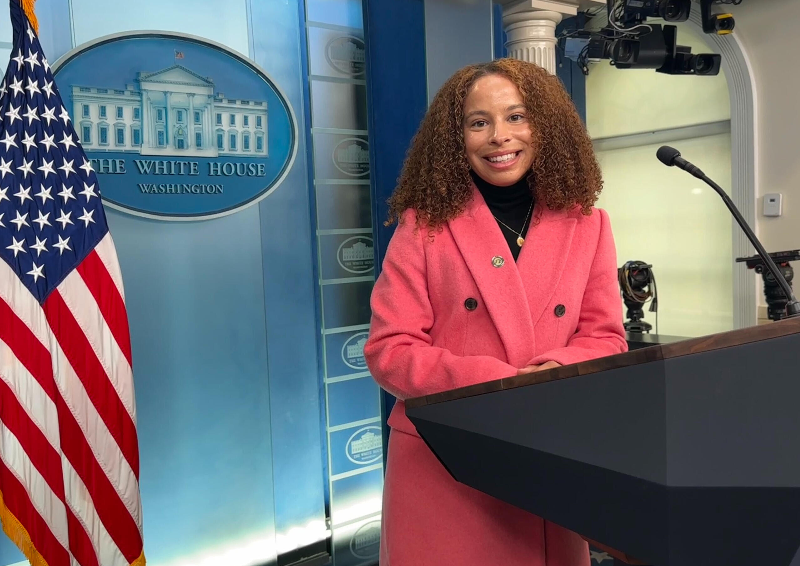 Avril Silva pictured in the White House Press Briefing room wearing a pink coat and black turtleneck.